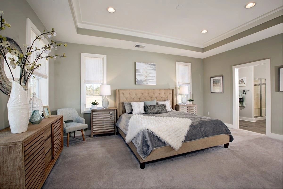 A serene model-home primary bedroom in soft sage and gray with a tufted upholstered bed and tray ceiling.